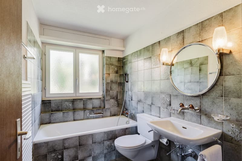 Modern bathroom with grey tiled walls, a bathtub, toilet, sink, and a large round mirror. Features a window with frosted glass and a wall-mounted light fixture.