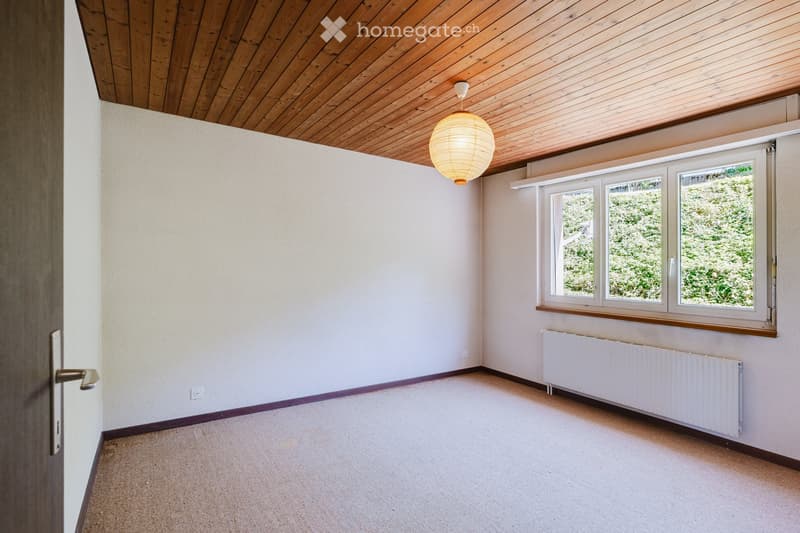 Empty room with light-colored walls, wooden ceiling, carpeted floor, large windows, radiator, and a hanging paper lantern light fixture.