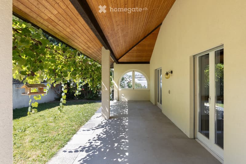 A covered outdoor corridor with a wooden ceiling, concrete floor, and glass doors leading to a grassy area with a bird feeder.