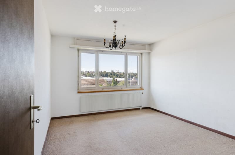 Empty room with white walls, carpeted floor, large window with view, radiator, and chandelier.