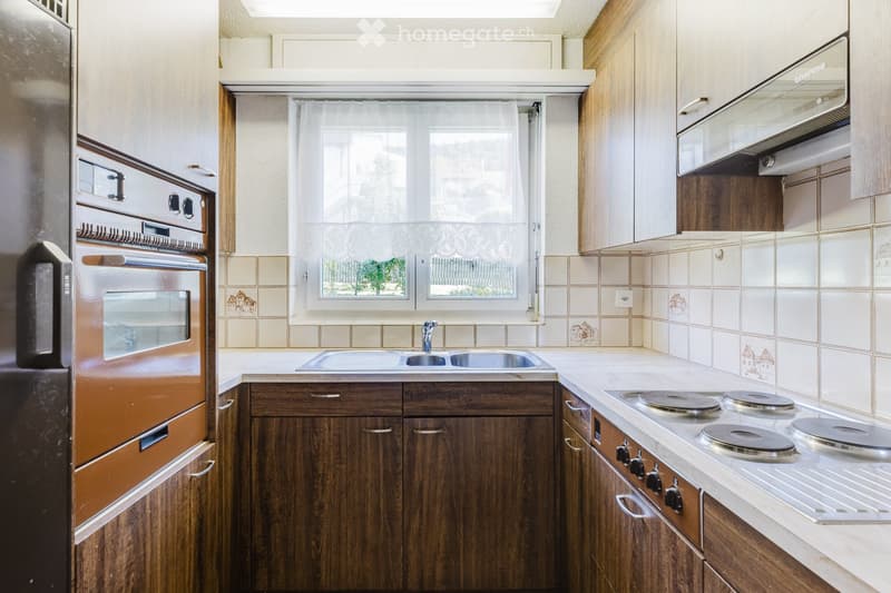 A well-equipped kitchen with wooden cabinets, a stainless steel sink, a four-burner electric stove, and a built-in oven. The space features tiled backsplash and a window with white curtains.