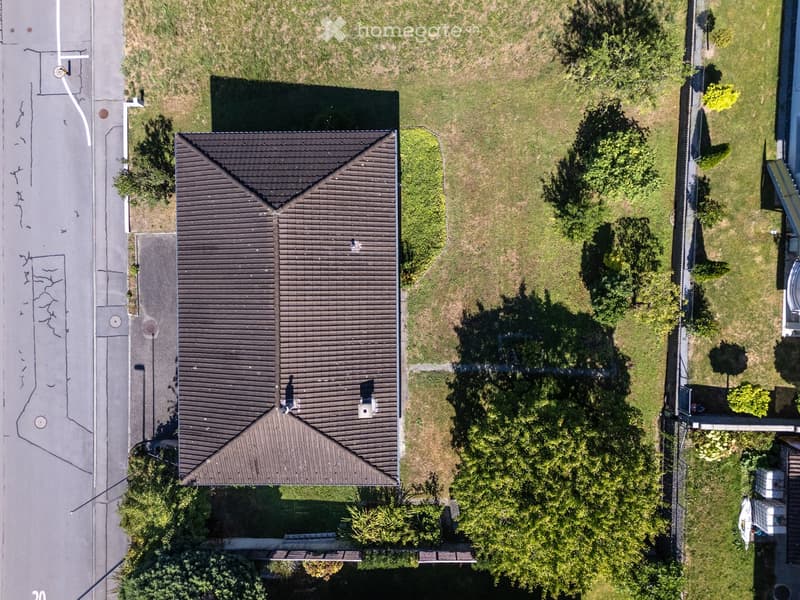 Aerial view of a two-story house with a dark roof, surrounded by a grassy yard and trees. The house has a chimney and is situated near a road with visible markings.