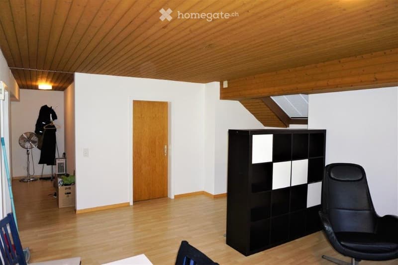 Bright, wooden-floored room with a black chair, a modern shelf unit, a small fan, and a coat rack with hanging clothes.