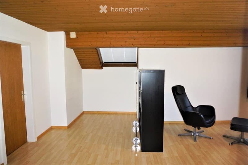 A modern, minimalist room with light wooden flooring and white walls. Features include a sleek black cabinet, a contemporary black chair with a matching stool, and a skylight window providing natural light.