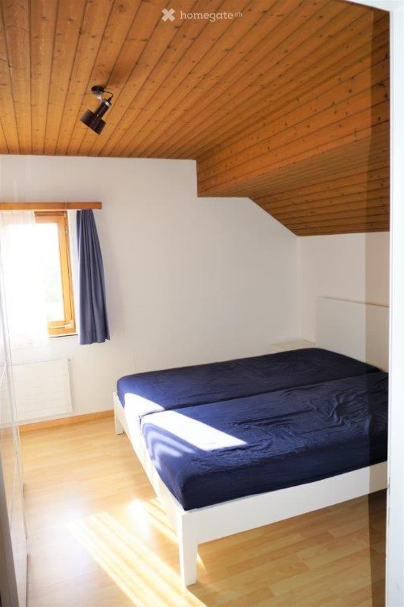 Cozy bedroom with a single bed, wooden floor, white walls, and a slanted wooden ceiling. Features a window with dark curtains and a wall-mounted light fixture.