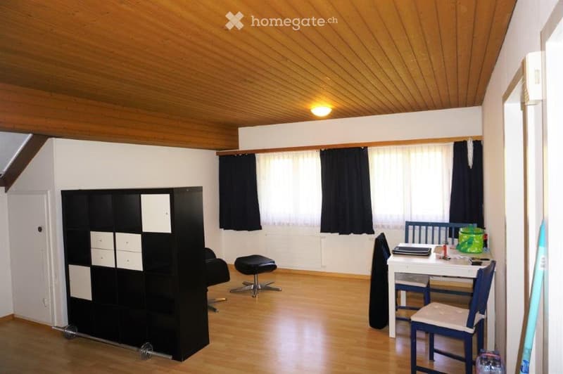 A furnished room with wooden flooring and a wooden ceiling. Features include a black wardrobe, a small stool, a chair, a table with a laptop, and a window with curtains.