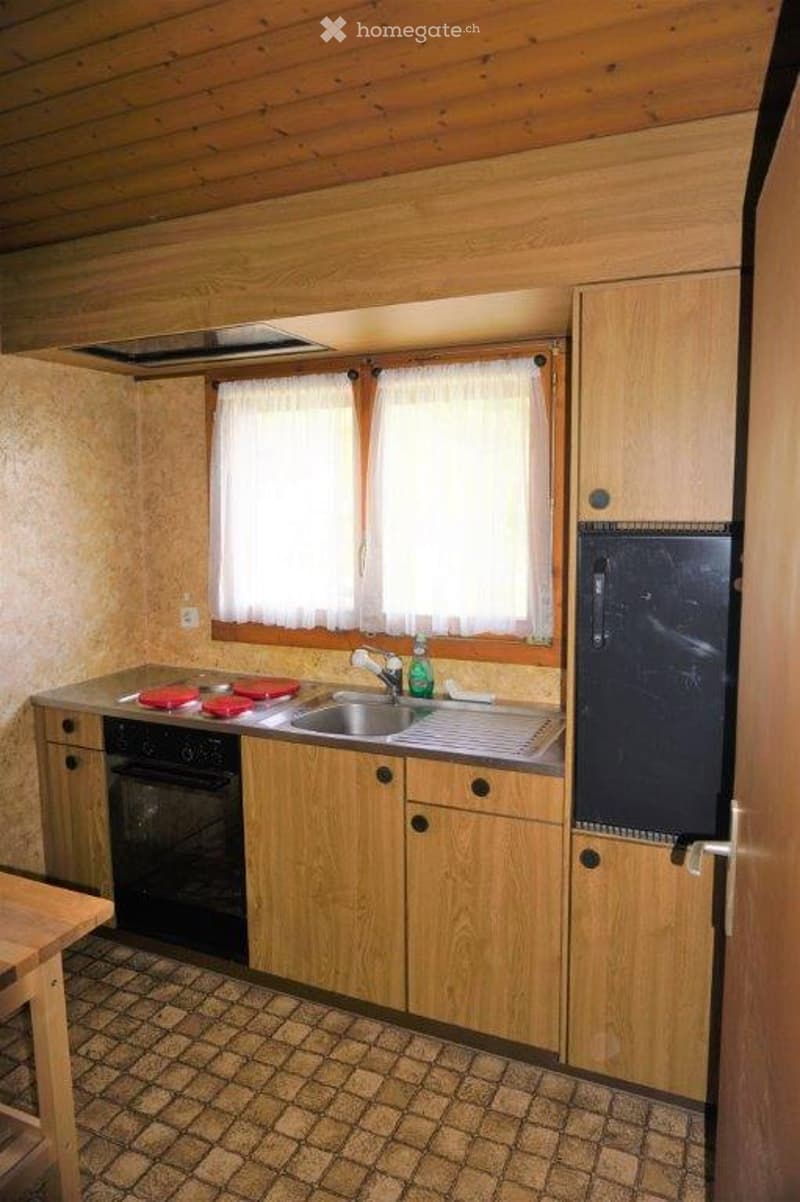 A cozy kitchen with wooden cabinets, a sink, an oven, and a refrigerator. The space features a tiled floor and a window with white curtains.
