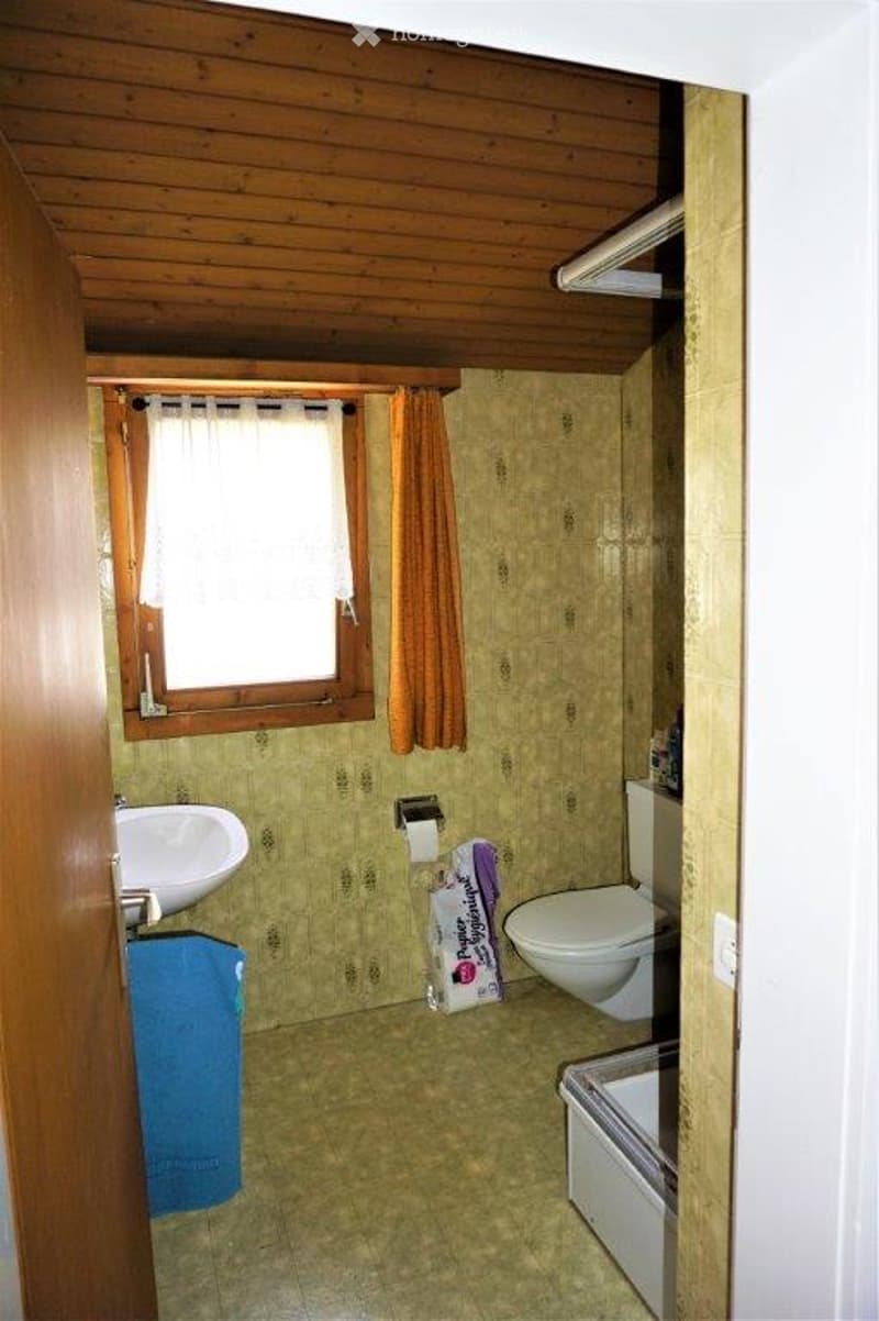 Bathroom with wooden ceiling, tiled walls and floor, a window with a curtain, a sink with a towel, a toilet, and a shower stall.