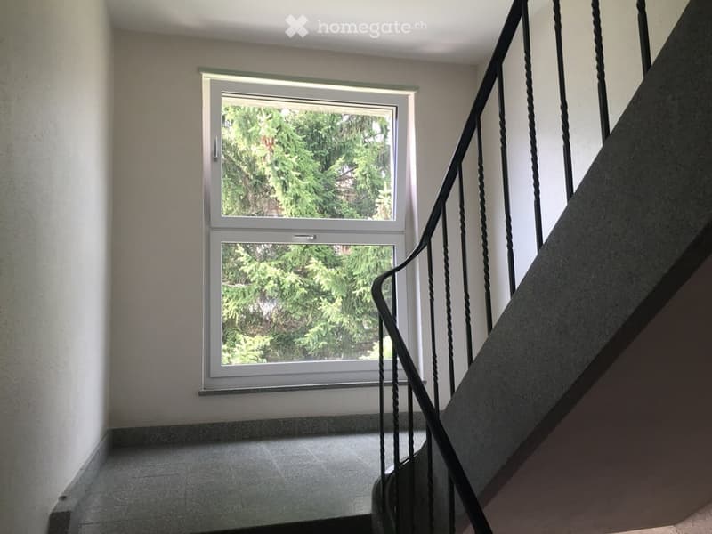 A modern hallway with a staircase featuring a black wrought-iron railing, a large window with a view of greenery, and tiled flooring.