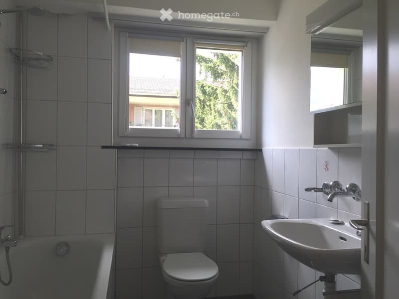 Modern bathroom with white tiles, a toilet, a sink, a bathtub, and a mirror above the sink. There is a window with blinds allowing natural light to enter.