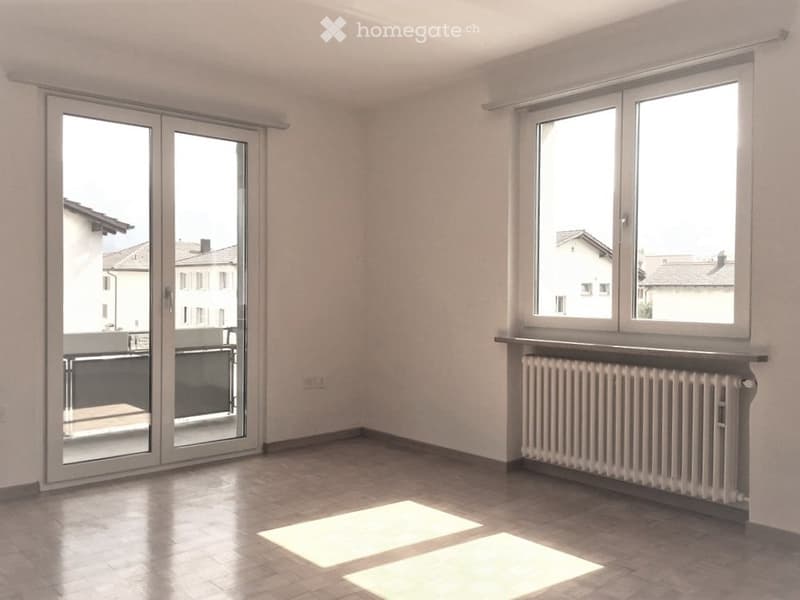 Empty room with tiled floor, large windows, sliding glass door leading to a balcony, radiator under the windows, and natural light streaming in.