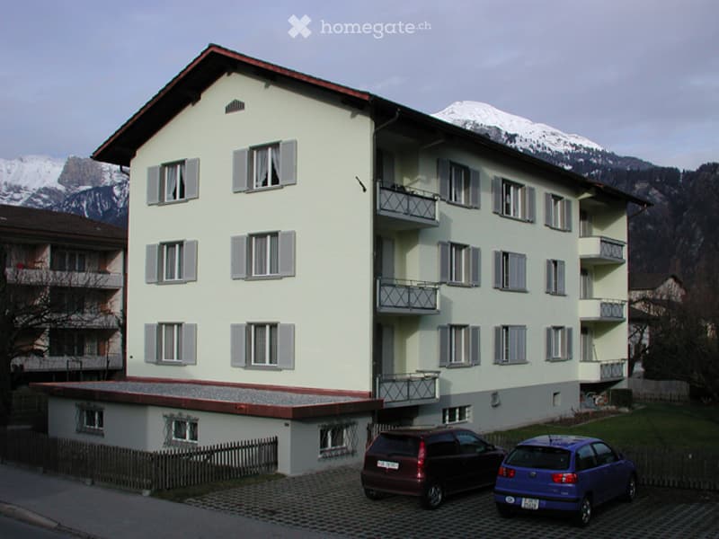 Multi-story residential building with light-colored facade, multiple windows, and balconies overlooking a mountainous landscape.