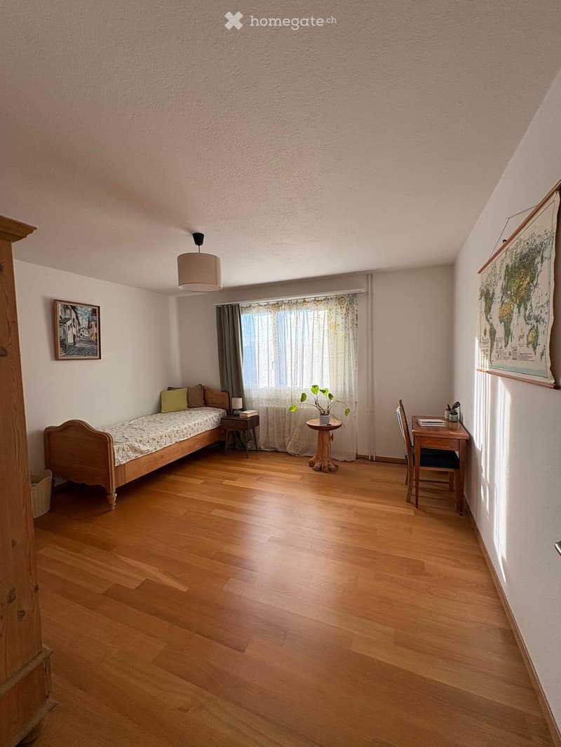 A small, well-lit room with wooden flooring, a single bed, a desk, a chair, a small table with a plant, and a lamp.
