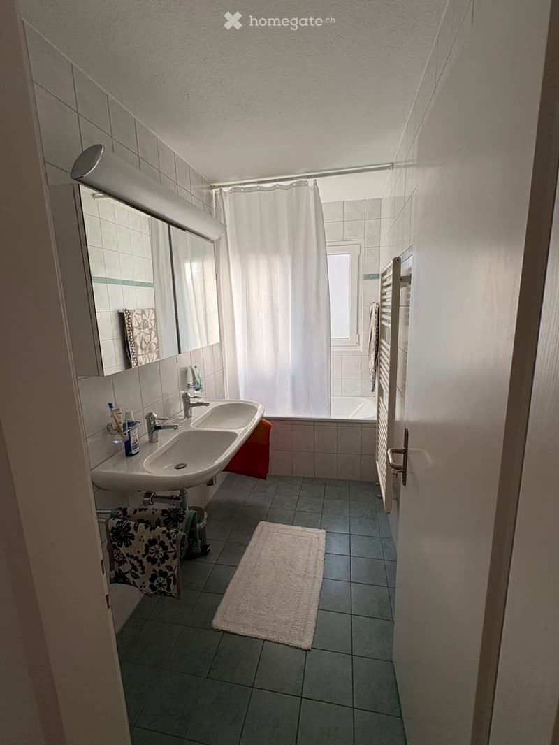 Modern bathroom with double sink, mirror, toothbrush holder, towels, bathrobe, and a shower area with a curtain. The floor is tiled, and there is a white rug in front of the sink.