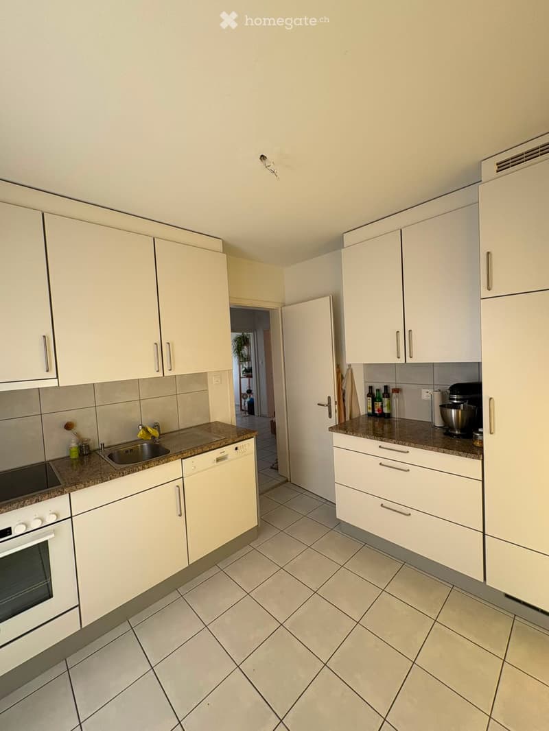 Modern kitchen with white cabinets, granite countertops, and stainless steel appliances including an oven, sink, dishwasher, and refrigerator. A mixer and several bottles are on the counter.