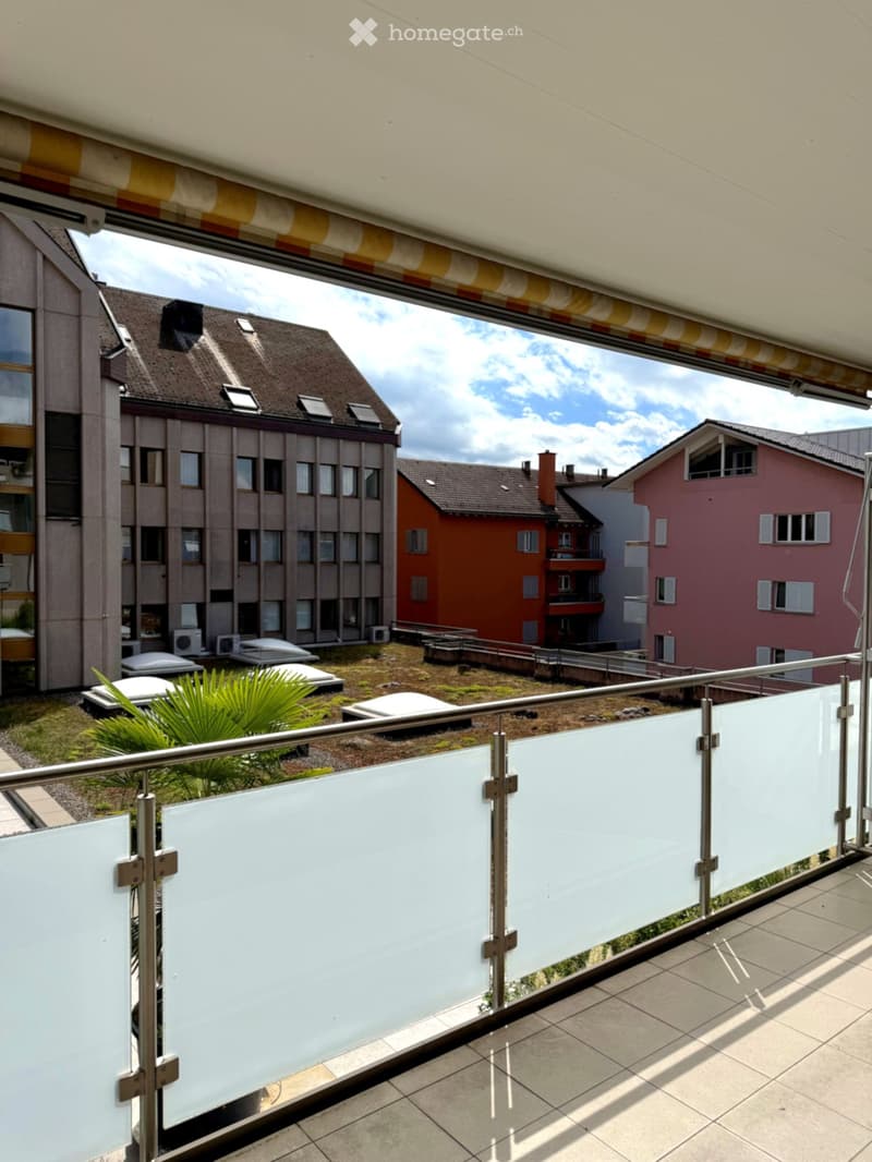 Balcony with glass railing, tiled floor, and view of surrounding buildings and greenery.