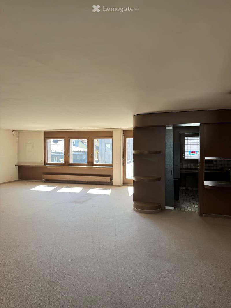 Empty room with beige carpet, large windows, and a small kitchen area visible in the background.