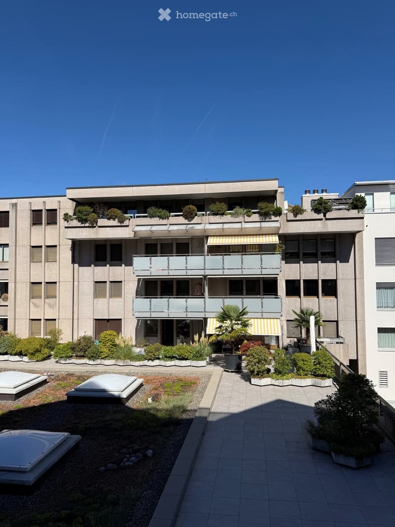 Modern multi-story building with balconies, greenery, and a landscaped courtyard area.