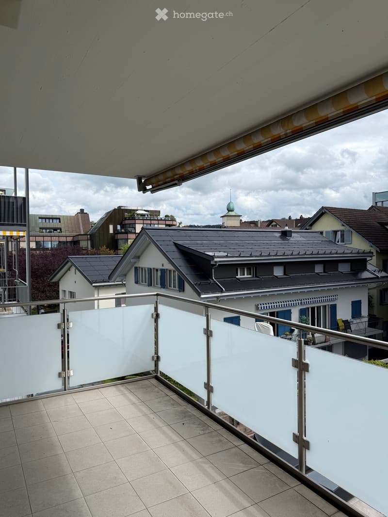Balcony with tiled floor, white glass balustrade, and metal railing, overlooking a residential area with houses and a cloudy sky.