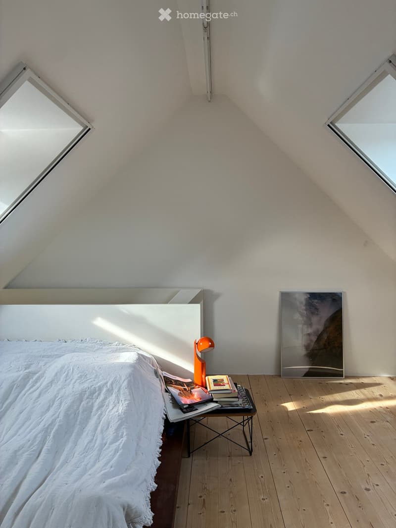 Bright, minimalist bedroom with wooden floors, white walls, and skylights. Features a white bed with a textured duvet, a small black nightstand with books and a lamp, and a large abstract picture frame on the wall.