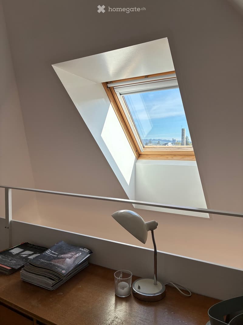 A cozy attic room with a slanted ceiling and skylight window, featuring a wooden table with a lamp, magazines, and a candle.