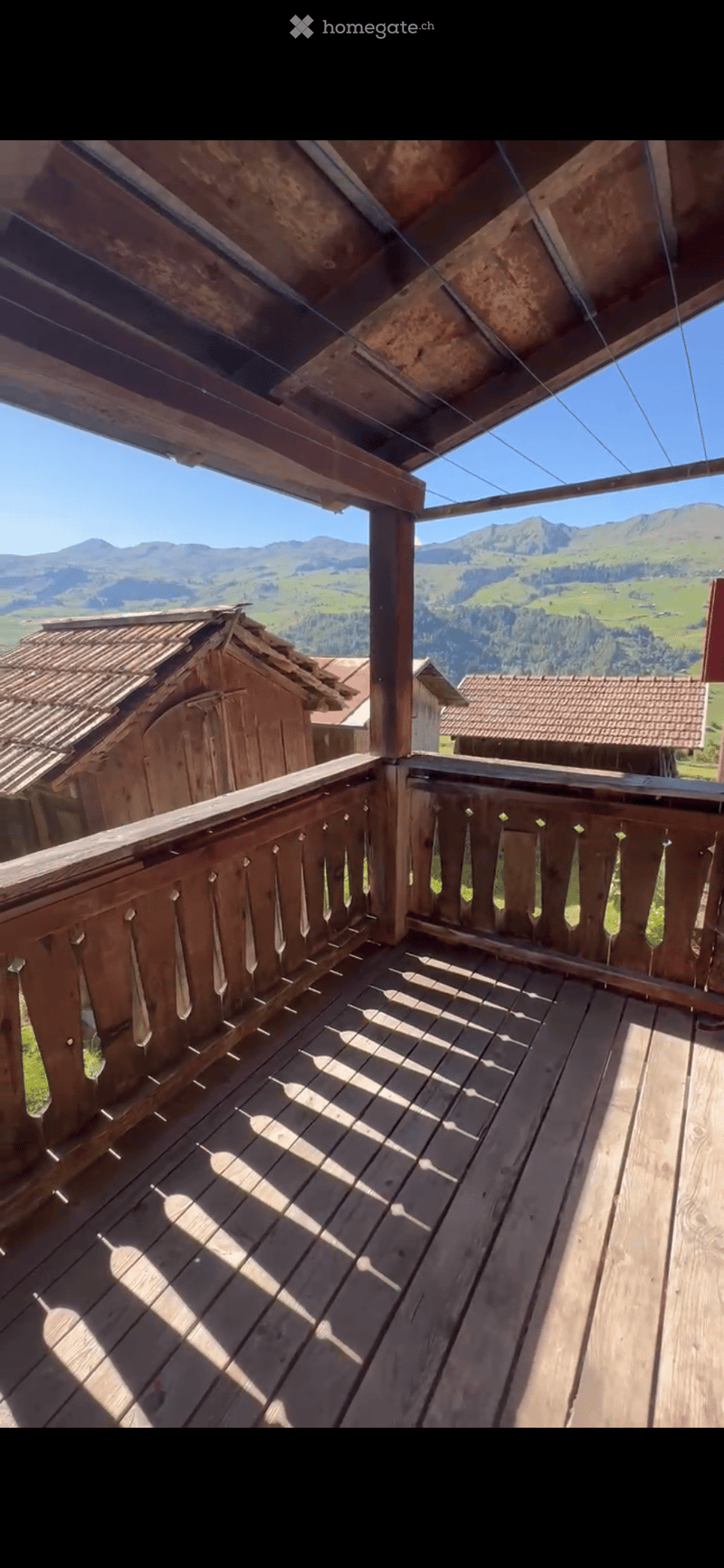 Wooden balcony with a railing, overlooking a scenic mountain landscape with green fields and scattered houses.