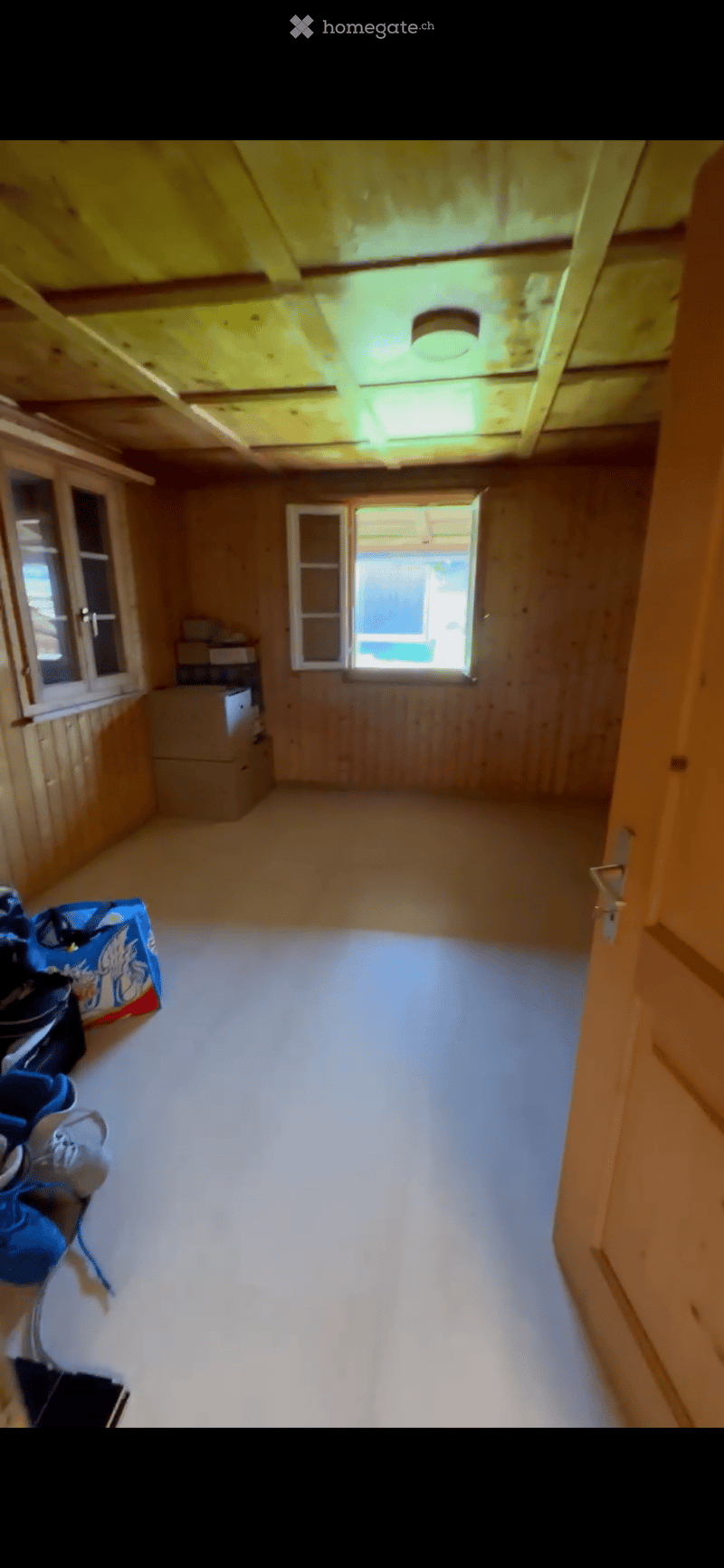 Empty room with wooden walls and ceiling, white floor, and a large open doorway leading outside. There are some stacked boxes and a few bags near the left wall.