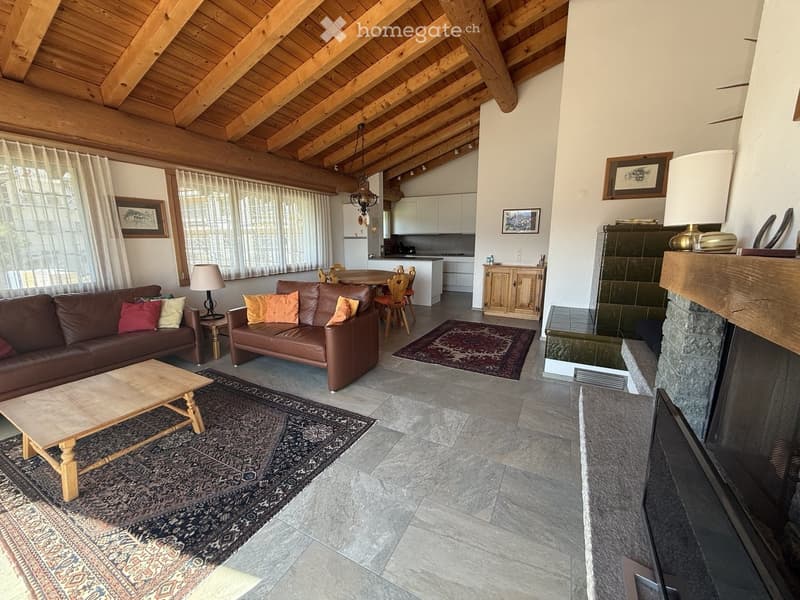 A spacious living room with exposed wooden beams, a stone fireplace, two leather sofas, a coffee table, a dining area with a table and chairs, and a modern kitchen in the background.