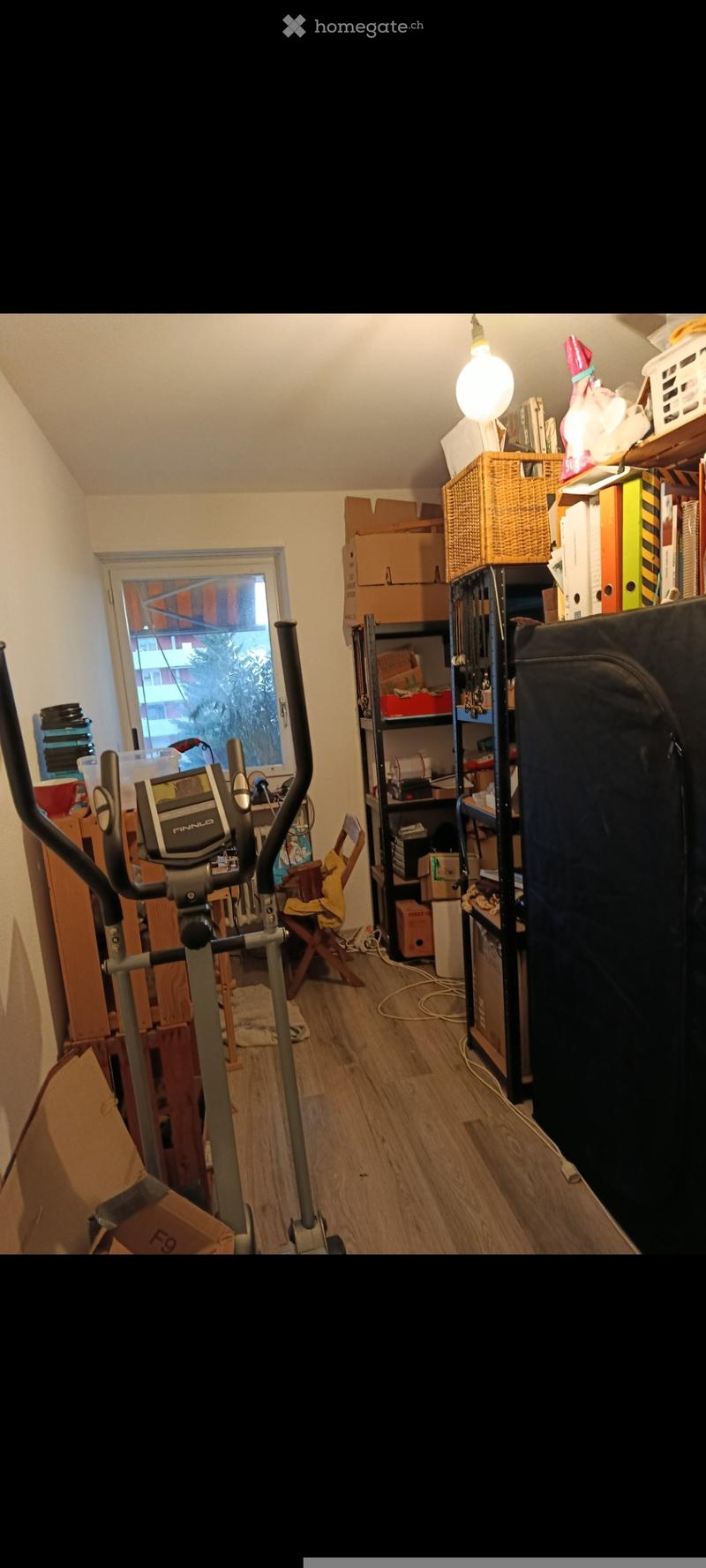 A cluttered room with wooden flooring, an exercise bike, shelves filled with books and storage bins, and several cardboard boxes scattered around.