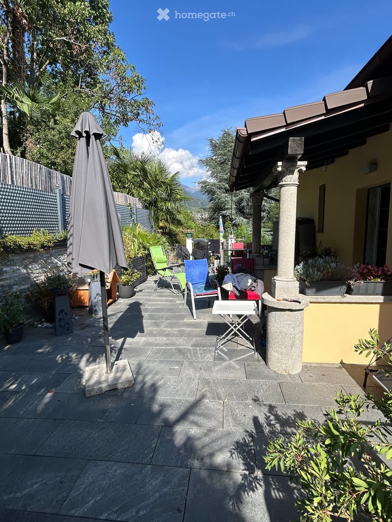 A well-maintained patio area with stone tiles, outdoor furniture including chairs and a table, potted plants, and an umbrella. The area is surrounded by greenery and has a view of mountains in the background.