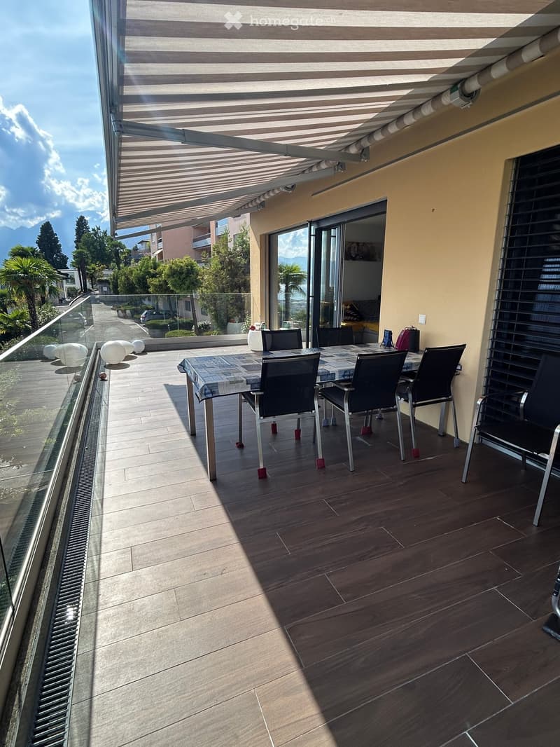 Balcony with wooden flooring, a table with chairs, and a retractable awning. The view includes trees and mountains.