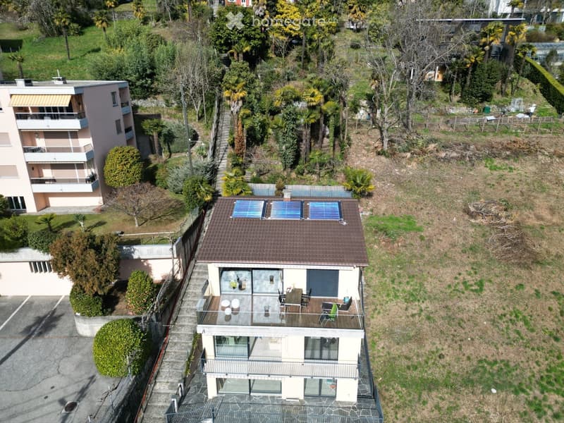 Aerial view of a two-story house with a brown tiled roof, solar panels, and a spacious balcony with outdoor furniture. The house is surrounded by greenery and trees, with a neighboring building visible to the left.