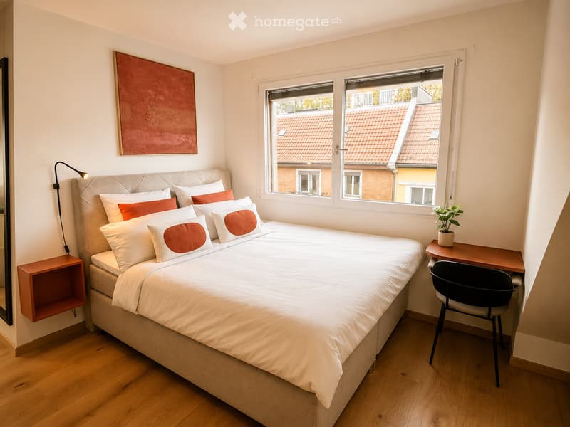Cozy bedroom with a large bed, white bedding, red accent pillows, a wall-mounted lamp, a small desk with a chair, and a potted plant on the desk. The room features wooden flooring and large windows with a view of rooftops.