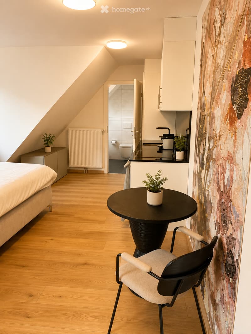 Cozy attic bedroom with wooden flooring, a bed, a small round table with a chair, a kitchen area with a counter, sink, and fridge, and a bathroom visible in the background.