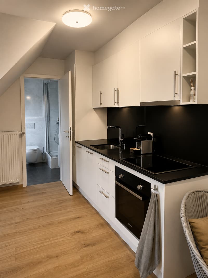 Modern kitchen with white cabinets, black countertops, and stainless steel appliances. Features a sink, faucet, oven, coffee maker, and a small chair with a cushion. A radiator is visible on the left wall, and a bathroom is partially visible through an op