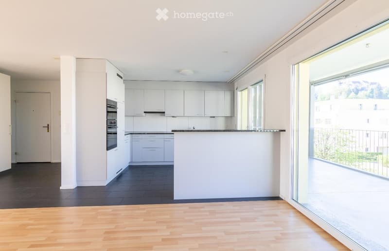 Modern kitchen with white cabinets, dark countertops, and a built-in oven. The space features a mix of dark tiled and light wooden flooring, with large windows offering a view of the outdoor area.