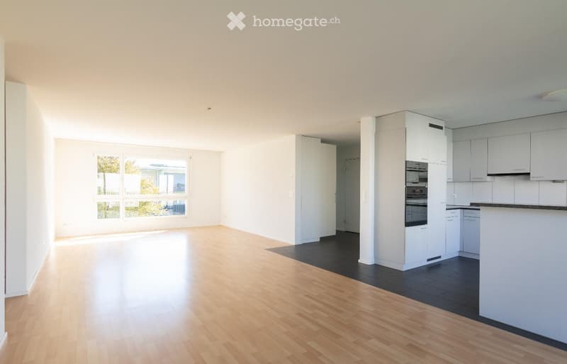 Empty room with light wooden flooring, white walls, and a small kitchen area with white cabinets and appliances.
