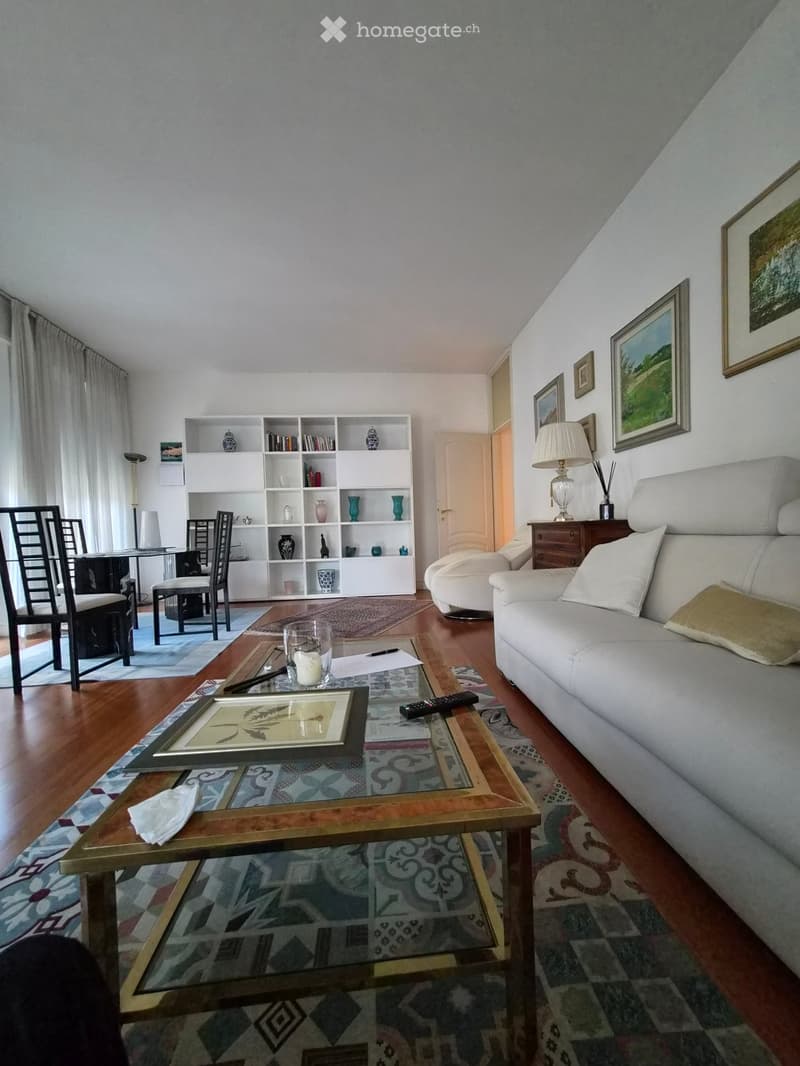 Living room with a white sofa, coffee table, dining area with chairs and table, bookshelf, and decorative items.