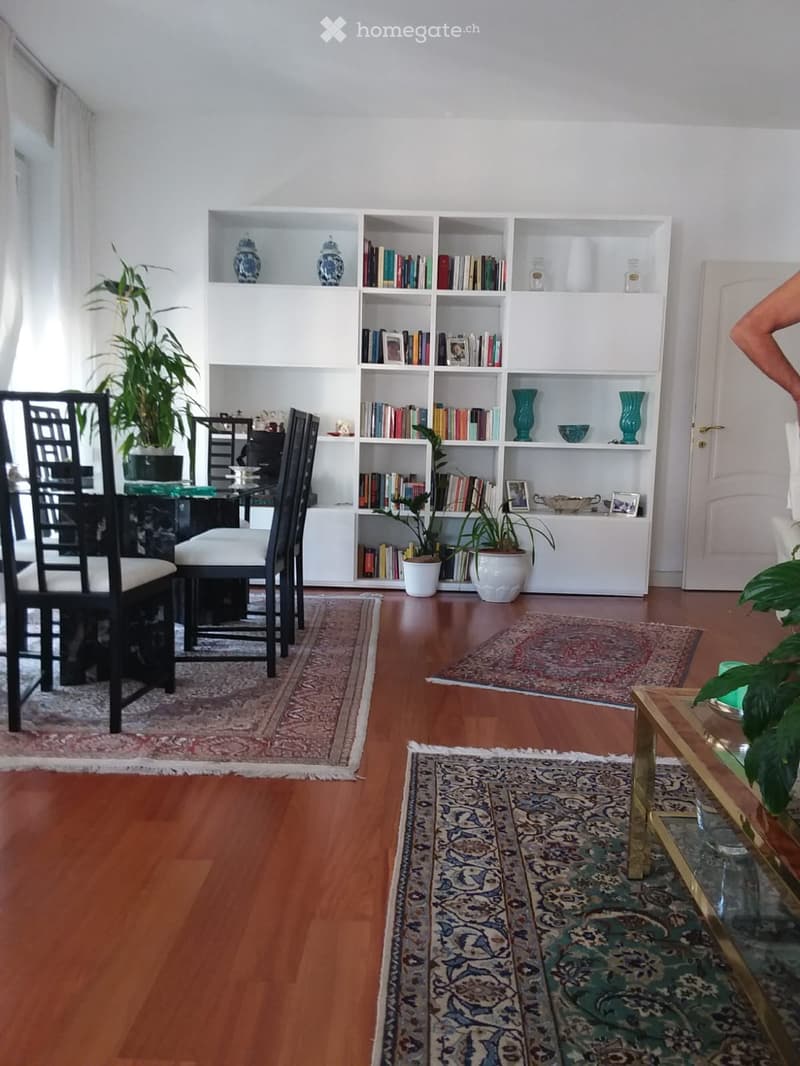 A well-lit living room with wooden flooring, multiple area rugs, a large bookshelf filled with books and decorative items, a dining area with chairs and a table, and a coffee table with a plant.
