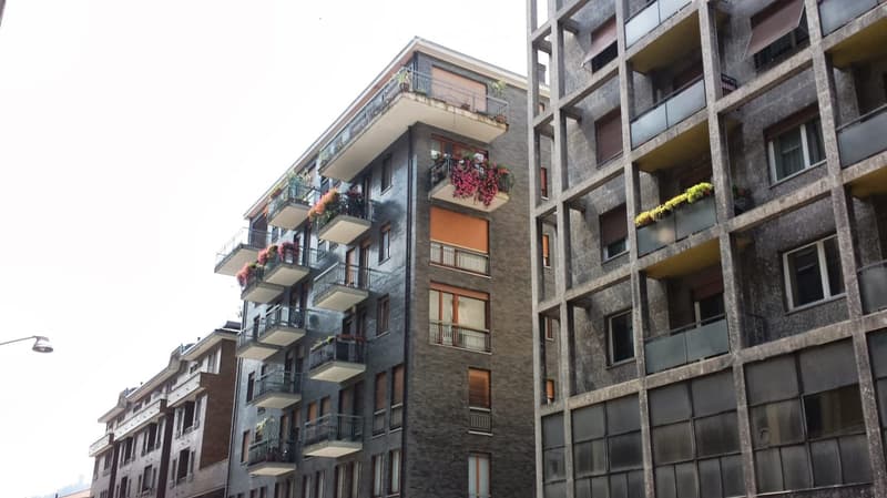 Modern multi-story apartment building with balconies and flower boxes, featuring a mix of large windows and smaller openings.
