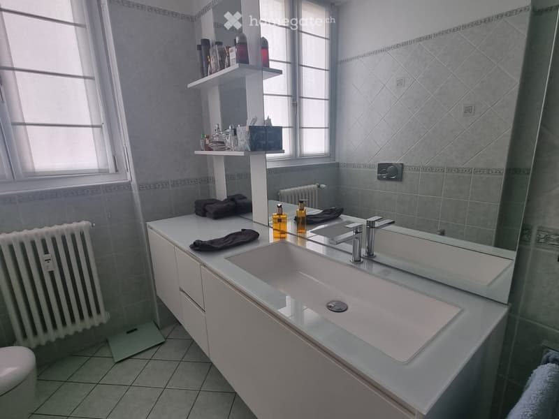 Modern bathroom with a double sink, large mirror, white cabinetry, and various toiletries on the counter. The room features tiled walls and a radiator.