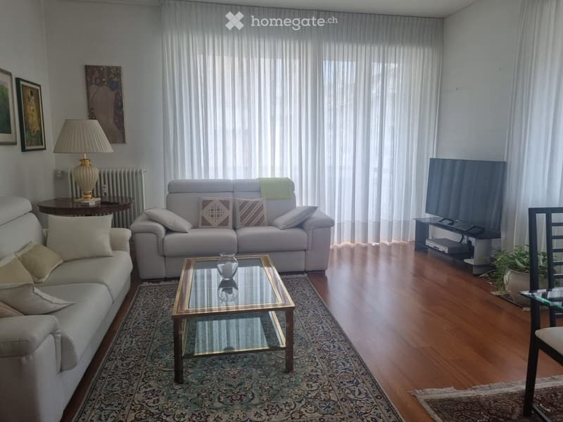 Living room with light-colored sofas, a glass coffee table, a table lamp, a television, and a patterned rug on a wooden floor.