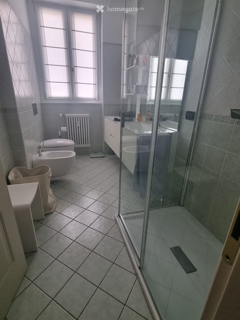 Modern bathroom with tiled floors, a toilet, bidet, bathtub, glass-enclosed shower, radiator, and a small stool near the toilet.