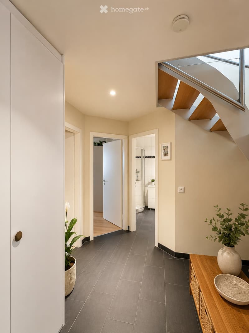 Modern hallway with grey tiled floor, white walls, and recessed lighting. Features a small wooden console table with a decorative bowl and a potted plant, leading to a bathroom and another room.
