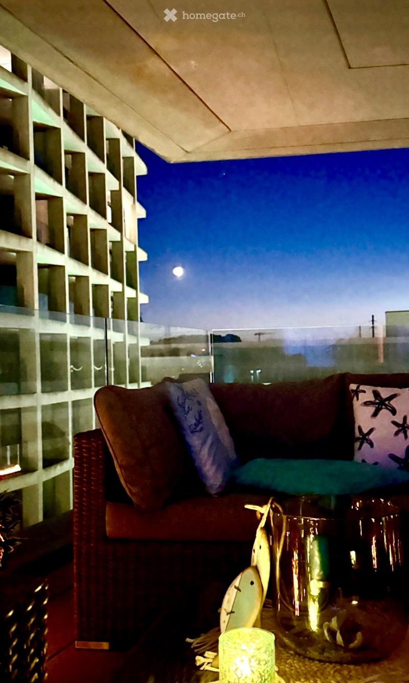 Balcony with a red sofa, decorative pillows, a small table with a vase and candle, and a view of a modern building at night.