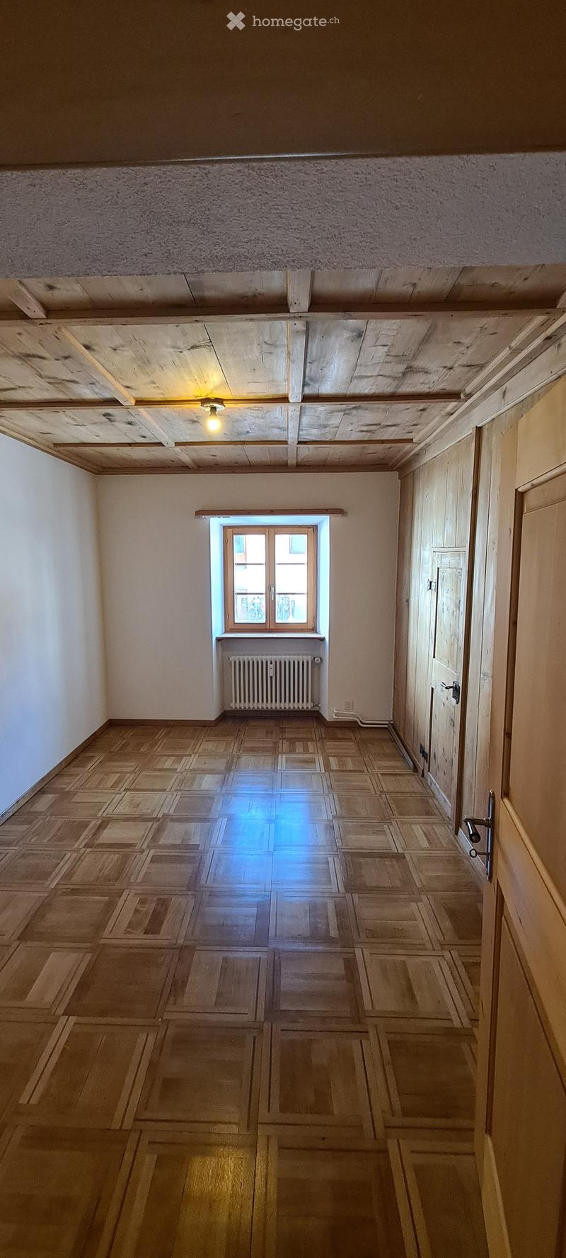 Empty hallway with wooden parquet flooring, white walls, exposed wooden beams on the ceiling, a single window with a radiator below, and two doors on the right side.