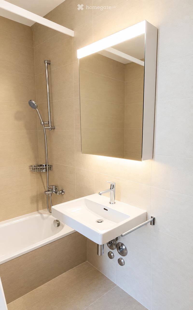 Modern bathroom with a white sink, bathtub, and a large mirror with built-in lighting. Features a sleek chrome faucet and a handheld showerhead with a rail.