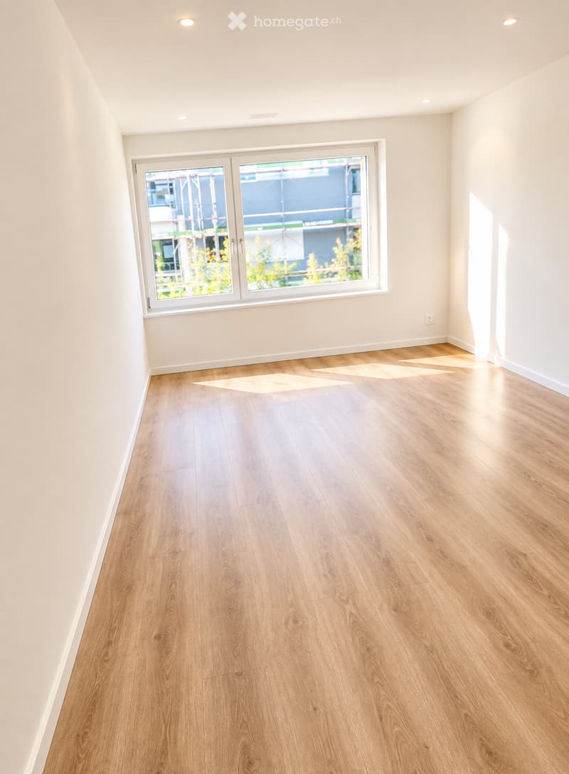 Empty room with light wooden flooring and white walls, featuring a large window with a view of the outside.