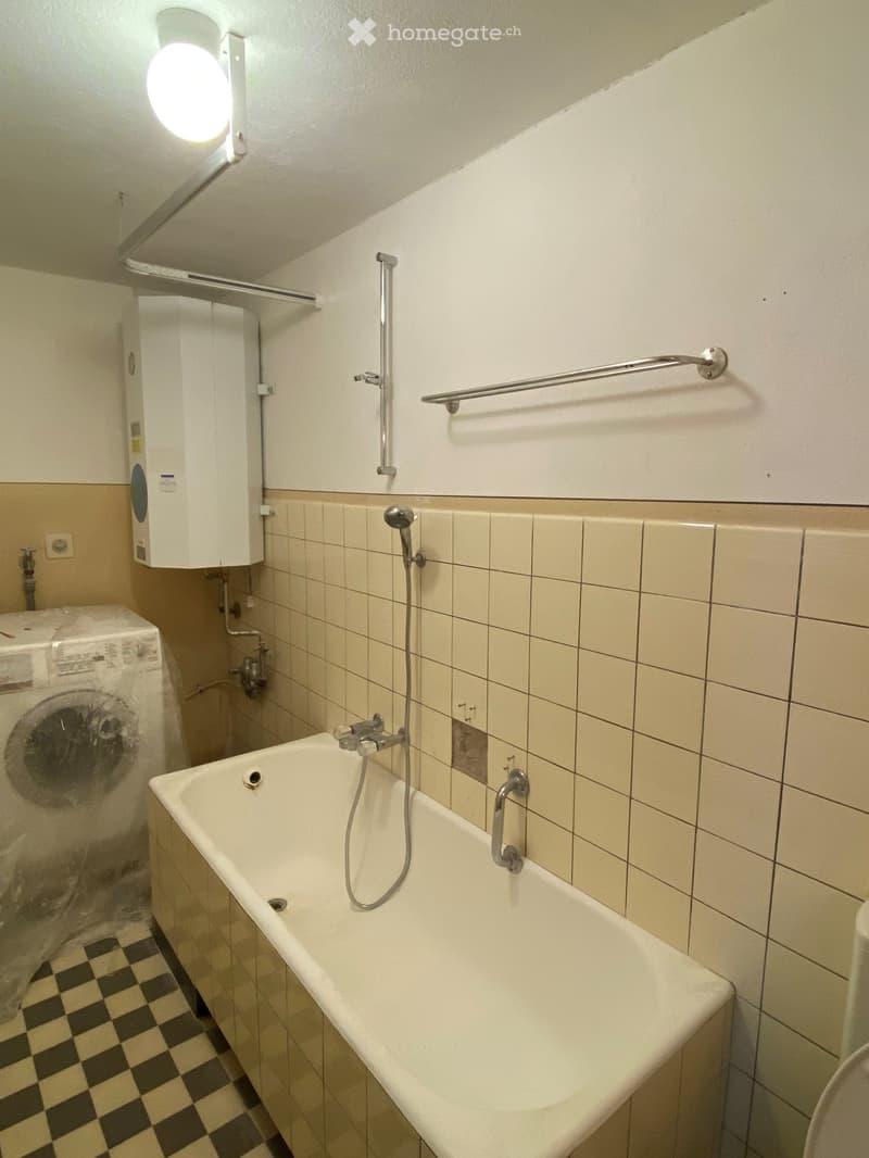 Bathroom with a bathtub, shower hose, handrail, washing machine, and water heater. The walls are tiled, and the floor has a checkered pattern.