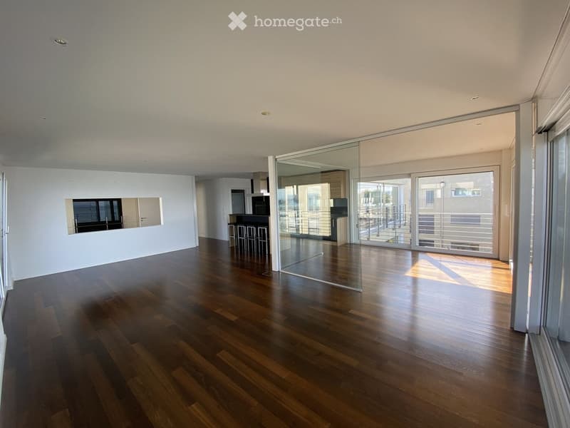 Empty room with wooden flooring, large glass sliding doors leading to a balcony, and a small seating area visible in the background.